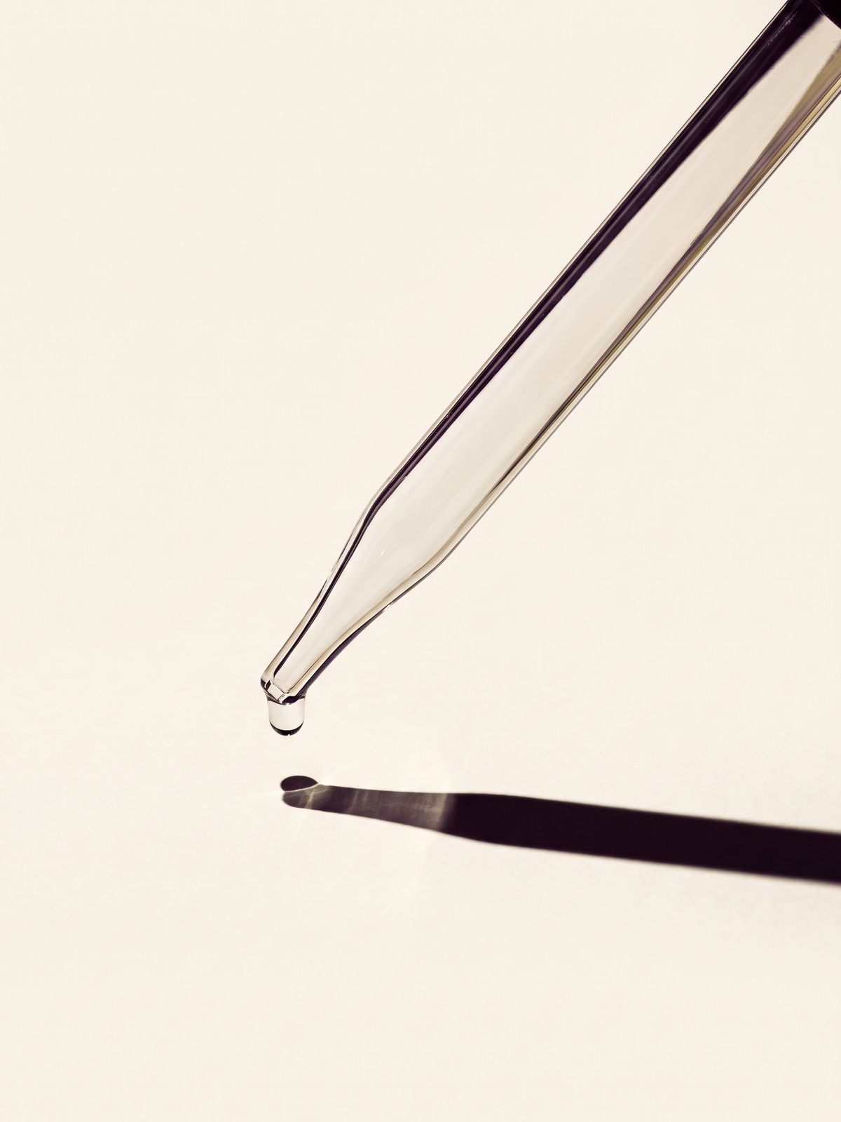 A glass pipette angled across a paper coloured surface with one drop held at its tip.